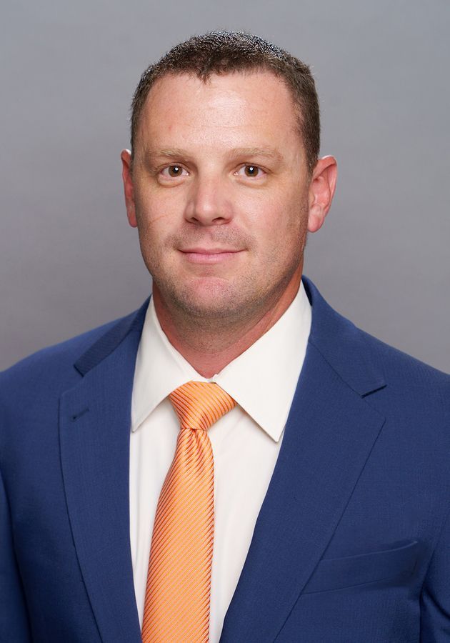 A man with short brown hair wearing a blue suit, white shirt, and orange tie against a gray background.