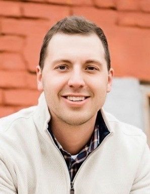 A smiling person wearing a cream-colored zip-up sweater over a plaid shirt, positioned in front of a brick wall.