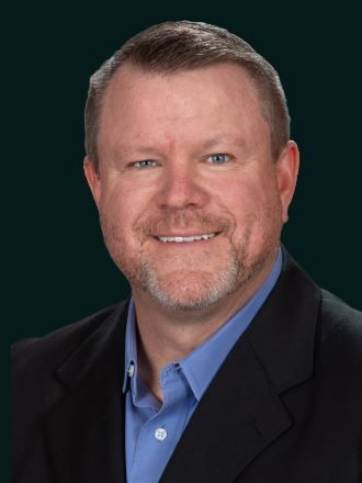 Headshot of a smiling person with a short beard, wearing a blue collared shirt and black blazer against a dark background.