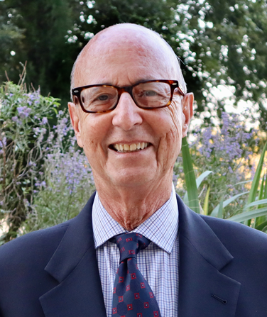 A smiling person with glasses, wearing a dark blue suit, patterned tie, and light blue checked shirt, outdoors.