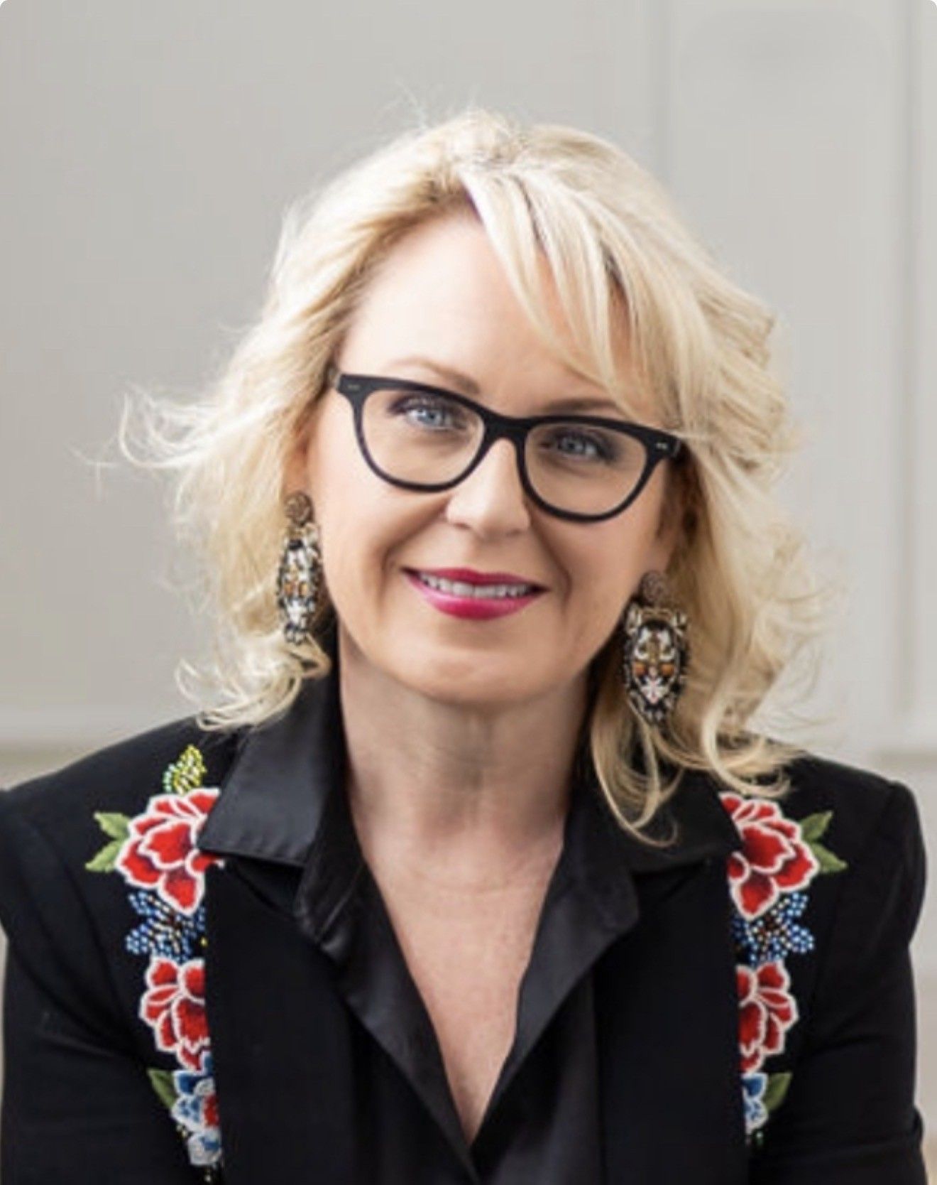A person with blonde hair and glasses wearing a black blazer with floral embroidery, smiling against a plain backdrop.