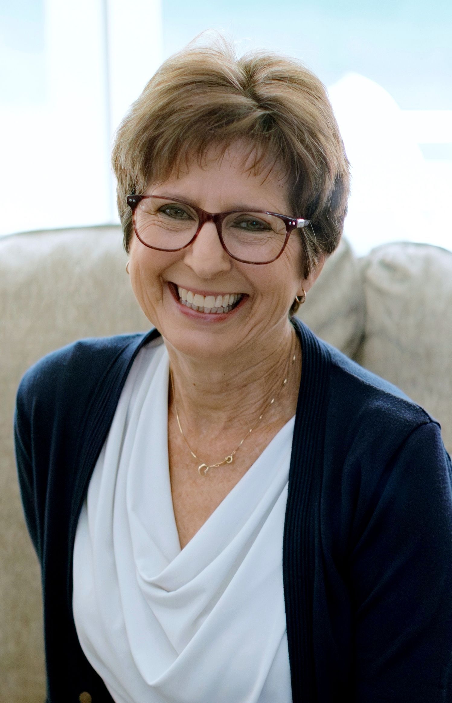 A smiling person wearing glasses, a white top, and a navy cardigan, sitting in front of a neutral-colored sofa.