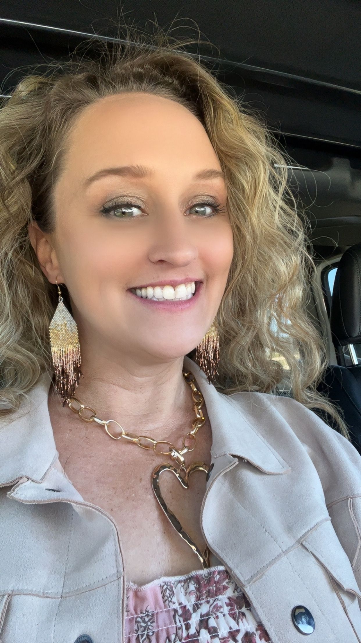 A smiling woman with curly hair, wearing a light jacket, a large heart-shaped necklace, and tiered beaded earrings.