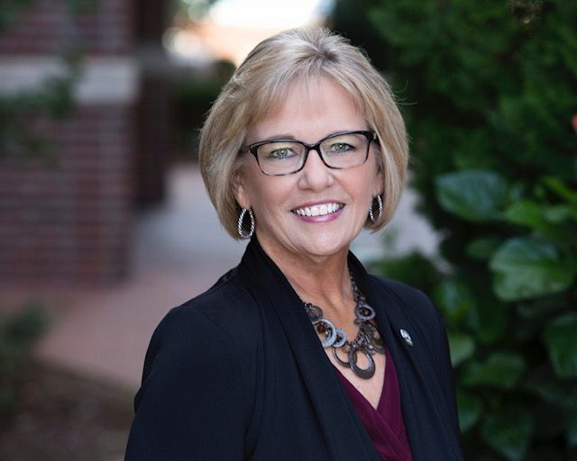 A professional headshot of a smiling person wearing glasses, a dark blazer, and a statement necklace outdoors.