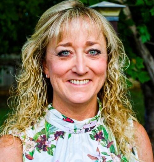 A smiling person with shoulder-length wavy blonde hair wearing a sleeveless white floral top, outdoors against greenery.