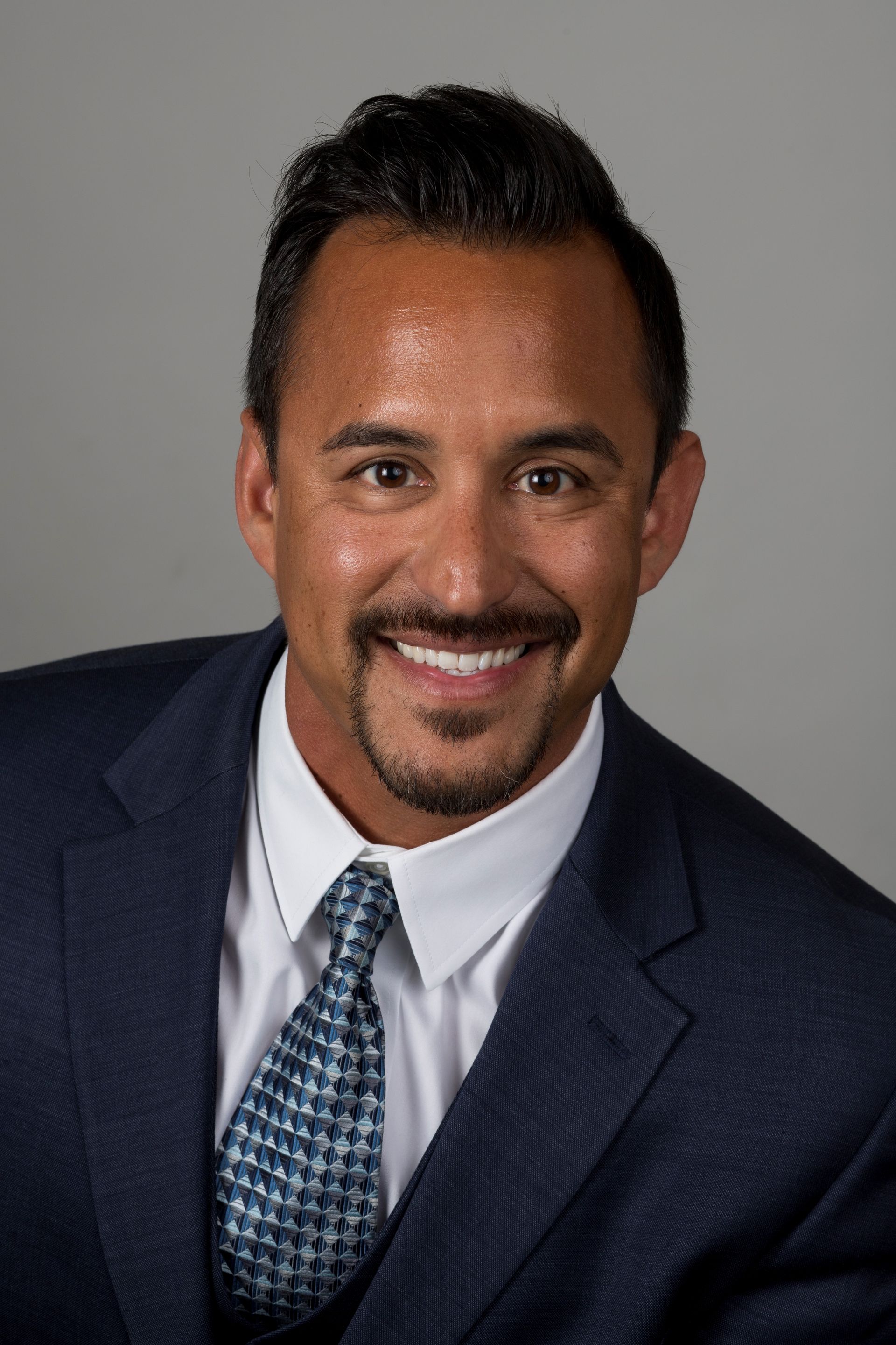Headshot of a smiling person wearing a dark blue suit, white collared shirt, and blue patterned tie against a gray backdrop.