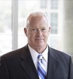 A man in a suit and striped tie smiles against a blurred, light-filled office background.