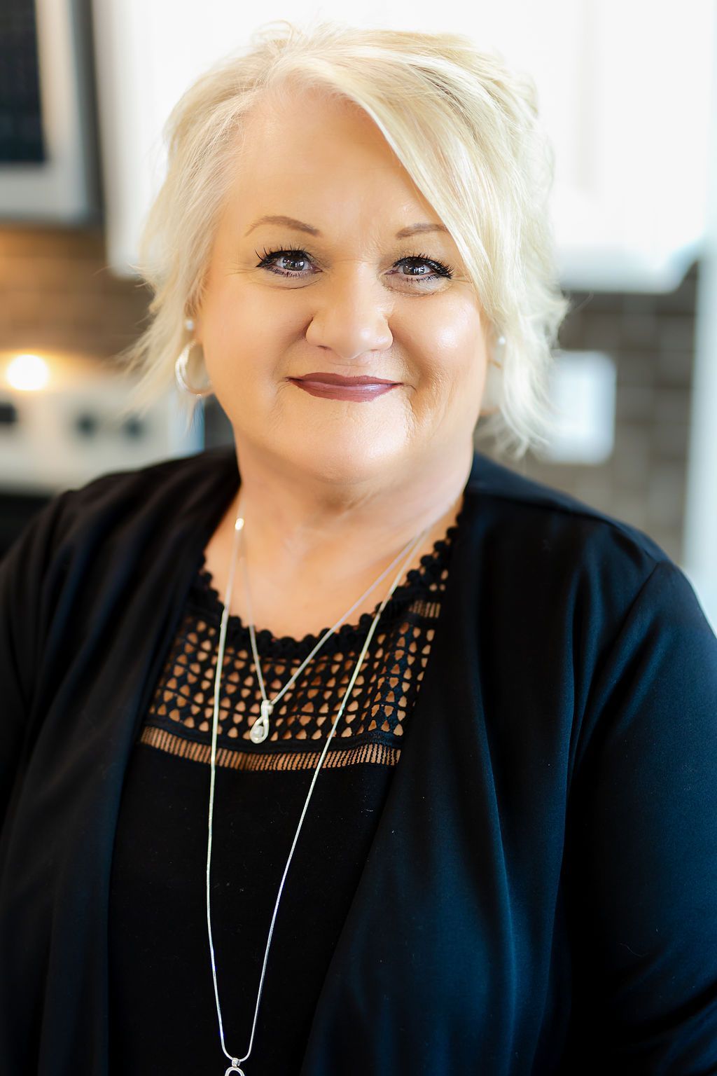 Headshot of a smiling person with blonde hair, wearing a black top with lace detail and a layered necklace.