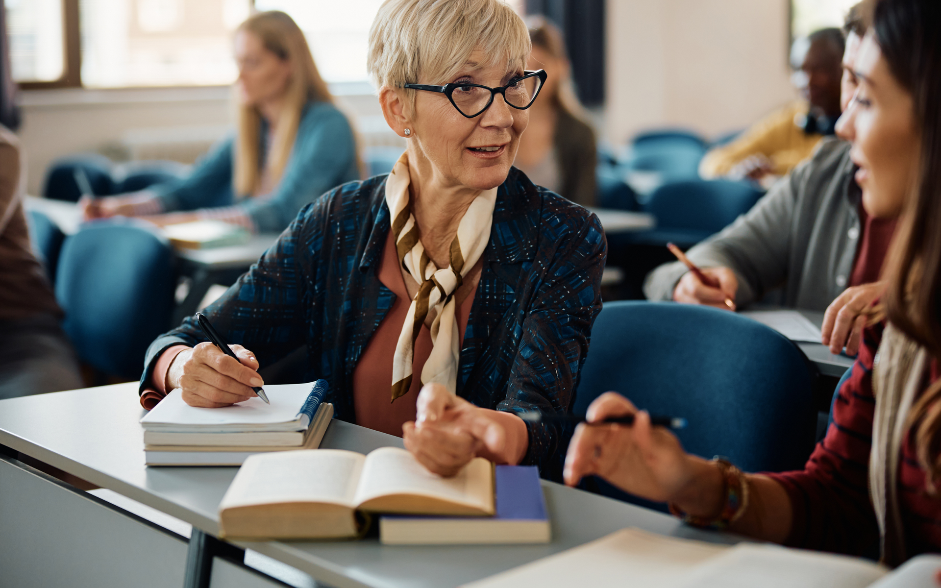 Students in a brightly lit classroom focused on their work, with one person writing in a notebook near an open book.