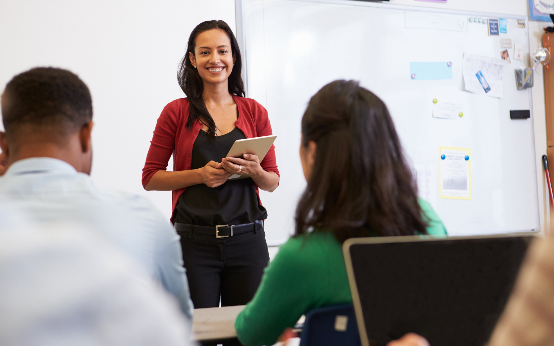A smiling instructor holding a tablet stands in front of a whiteboard in a classroom, facing students.