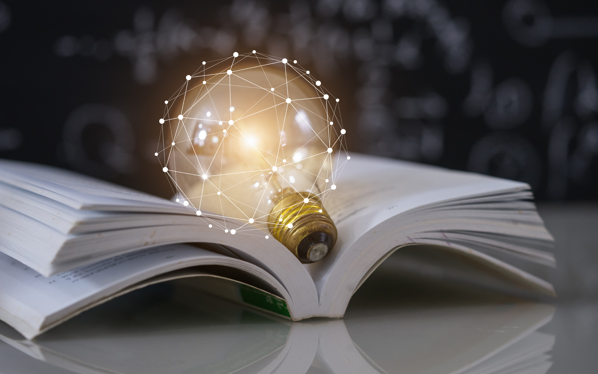 A glowing light bulb with a digital network pattern rests on an open book against a dark, chalk-covered background.