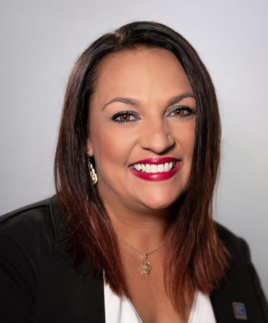 A professional headshot of a smiling person with dark hair, wearing a black blazer over a white top and a gold necklace.