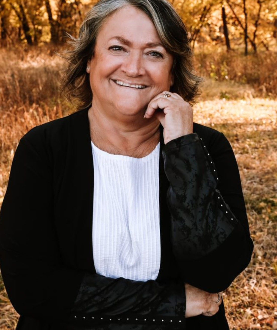 Smiling person with grey-highlighted hair in a black cardigan and white top, posing outdoors in a wooded setting.