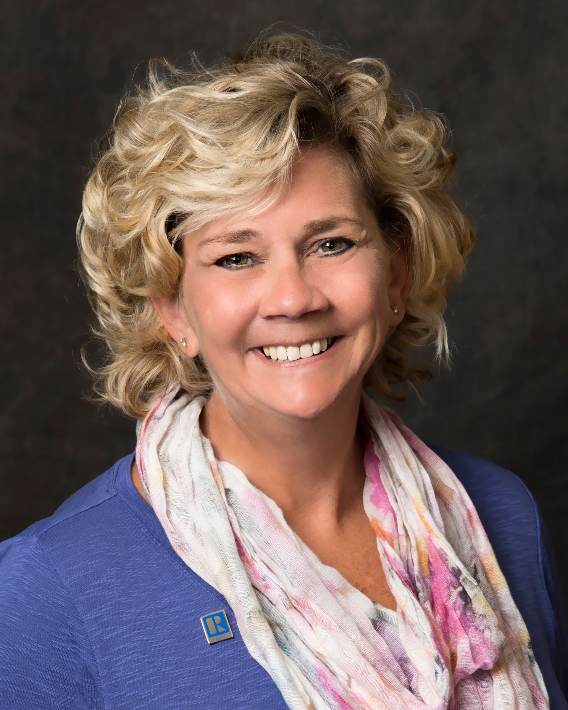 A smiling woman with curly blonde hair wearing a blue top and a light-colored patterned scarf against a dark background.