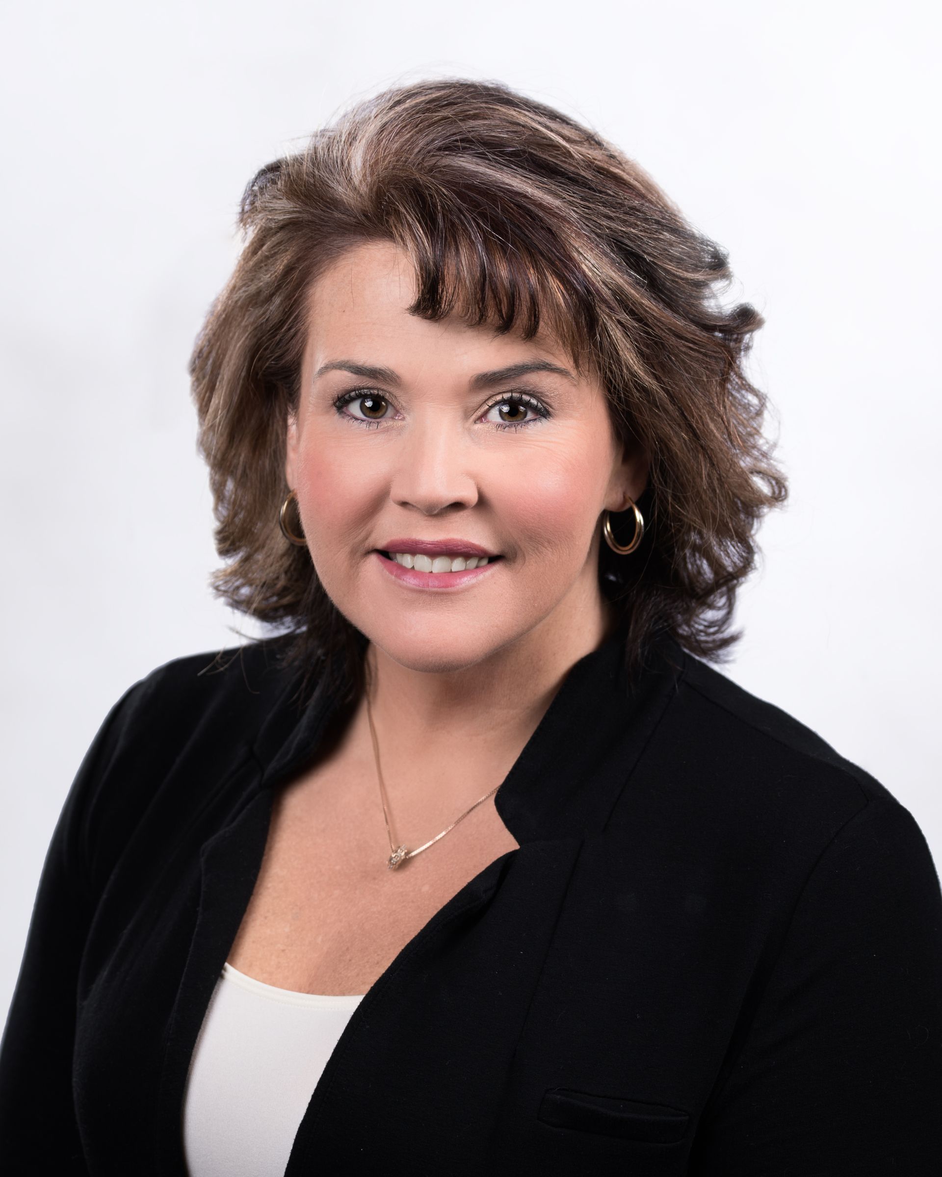 Professional headshot of a woman with shoulder-length brown hair, smiling, wearing a black blazer against a white background.
