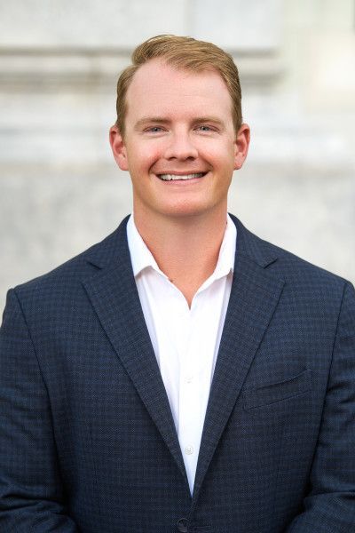 A headshot of a person smiling, wearing a dark blue patterned blazer and a white collared shirt against a light background.