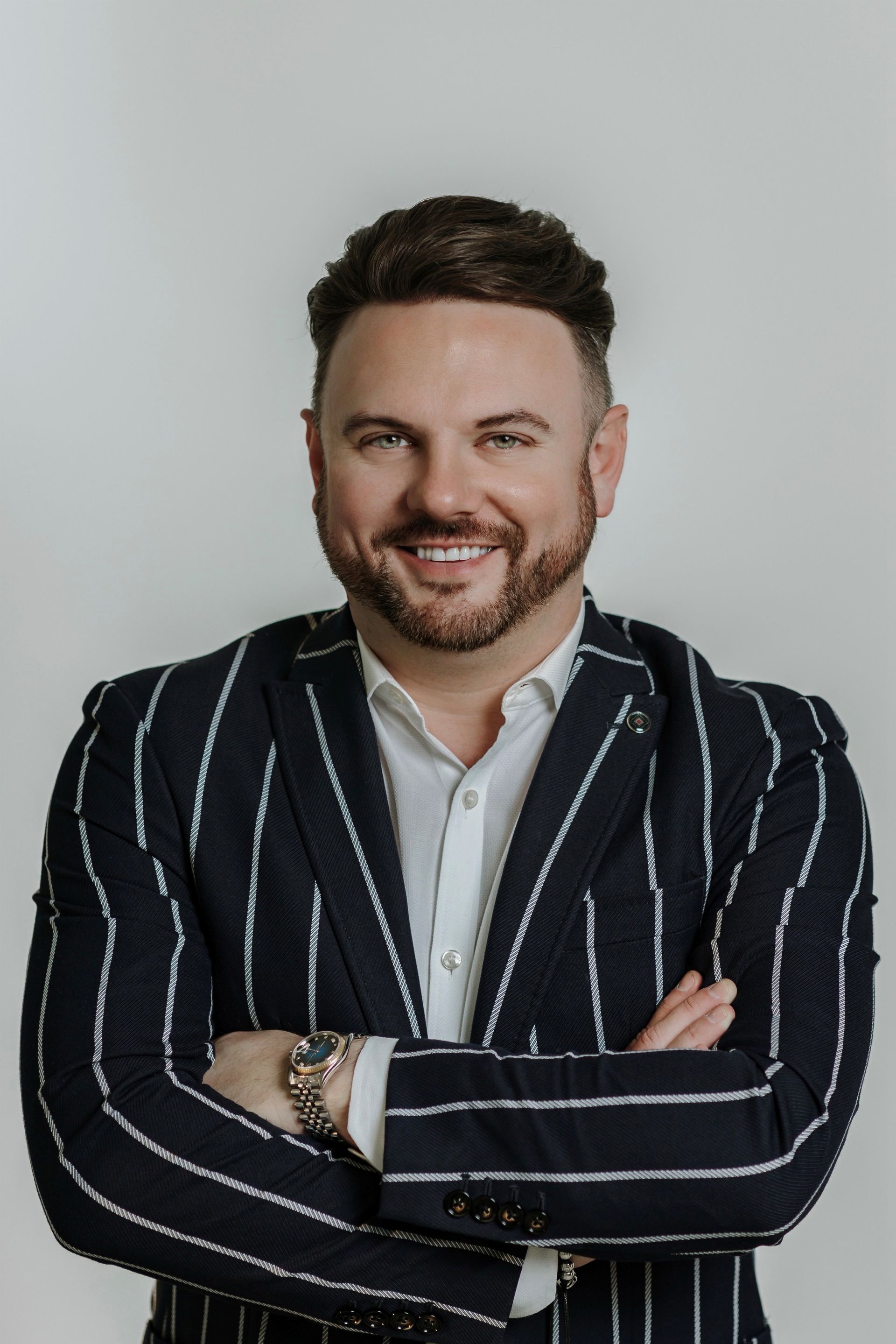A smiling person with a beard, wearing a white shirt and a navy blue pinstriped blazer, with arms crossed against a wall.