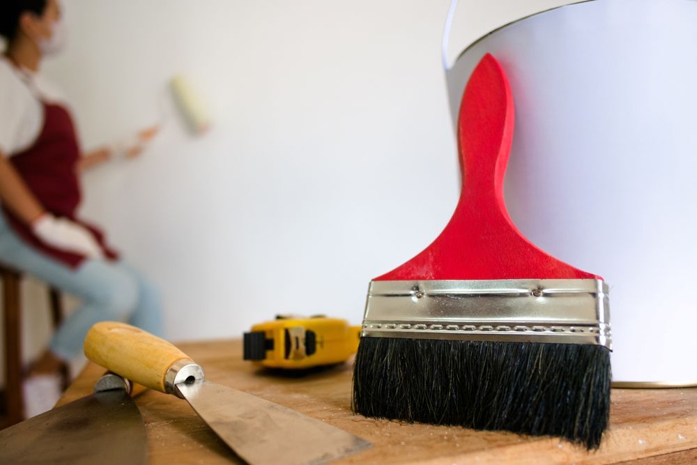 A Red Paintbrush Leaning Against A Bucket With A Woman Painting In The Background — L & S Wirth Painting in Dubbo, NSW