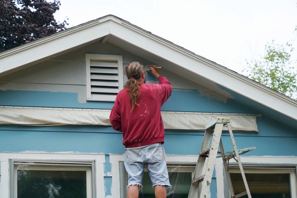 A Man Is Standing On A Ladder Painting A Blue House — L & S Wirth Painting in Narromine, NSW