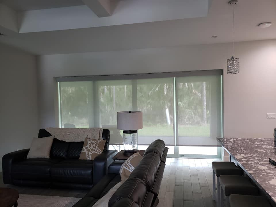 White vertical blinds covering a large window; a glimpse of outside visible through a partially open door.