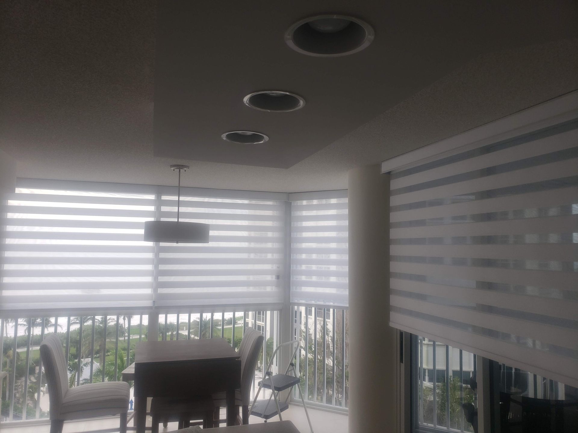 Interior view of a balcony with white zebra blinds, dining table and chairs, recessed ceiling lights.