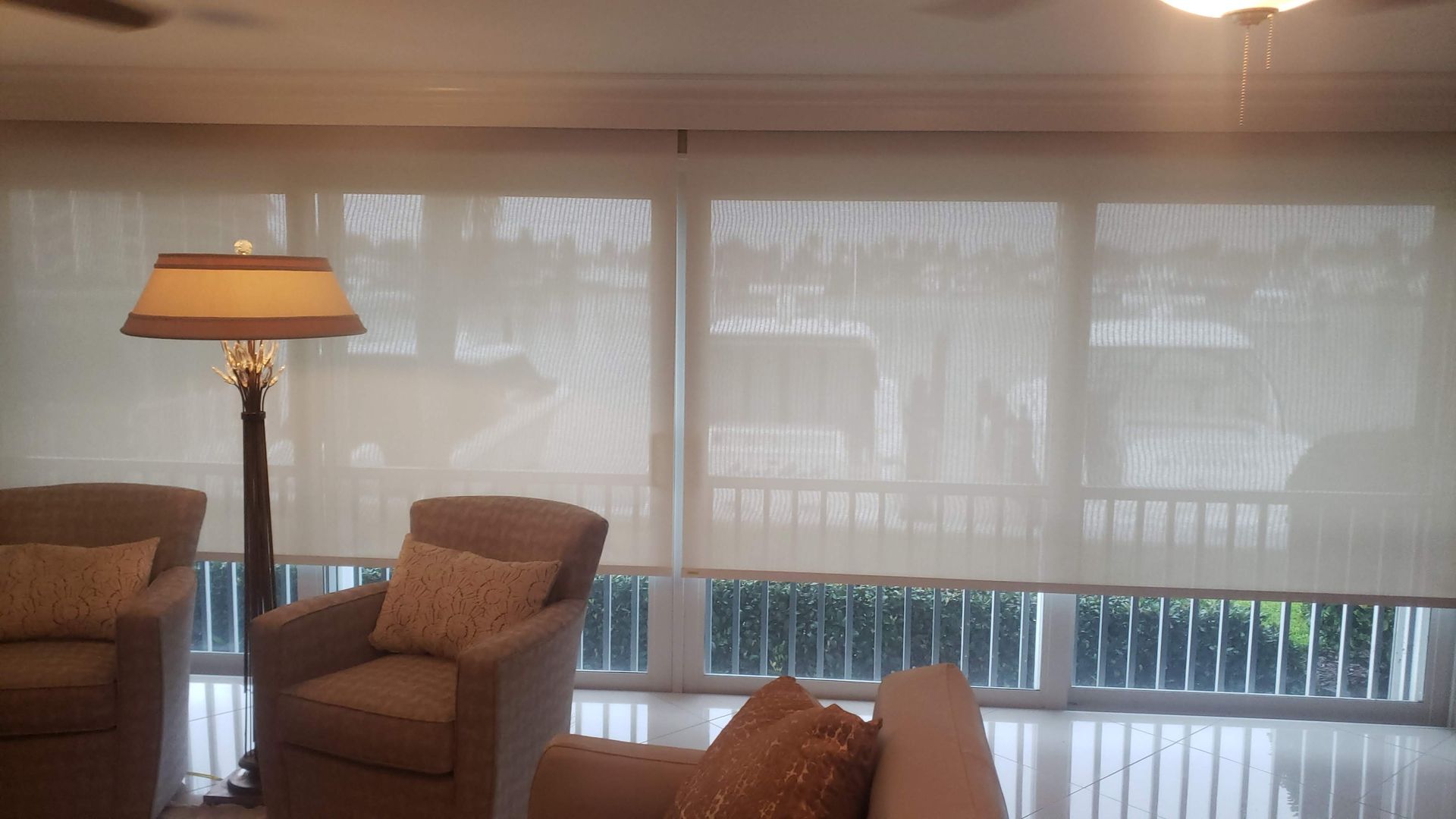 Living room with beige roller shades covering large windows. Beige armchairs, floor lamp, and outside greenery visible.
