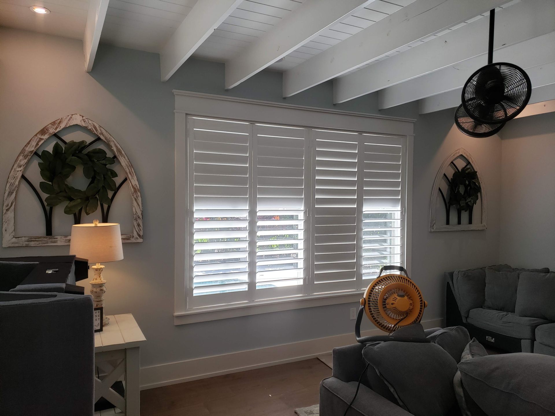 Living room with white shutters, gray couch, wooden beams, and arched wall decor.