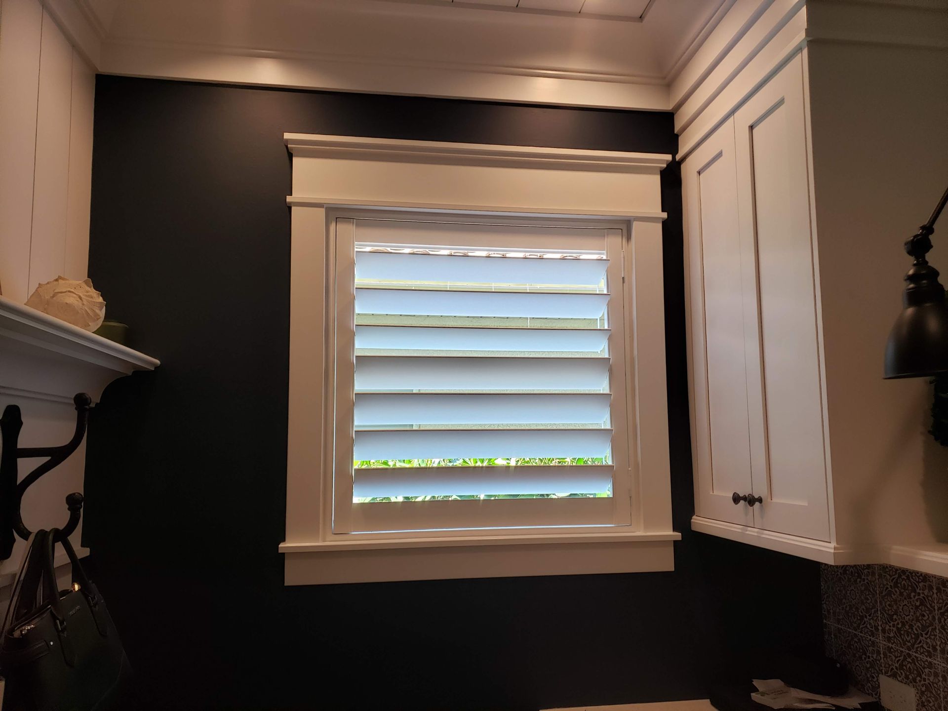 White shutters in a kitchen window, surrounded by dark blue wall. White cabinetry on the right.