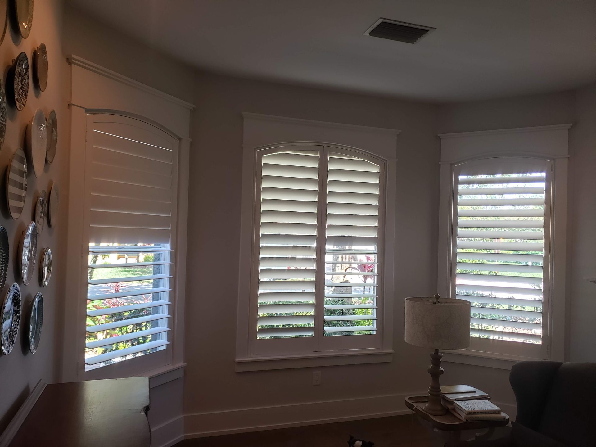 Bay windows with white shutters and trim. A lamp sits on a table. Decorated plates on the wall.
