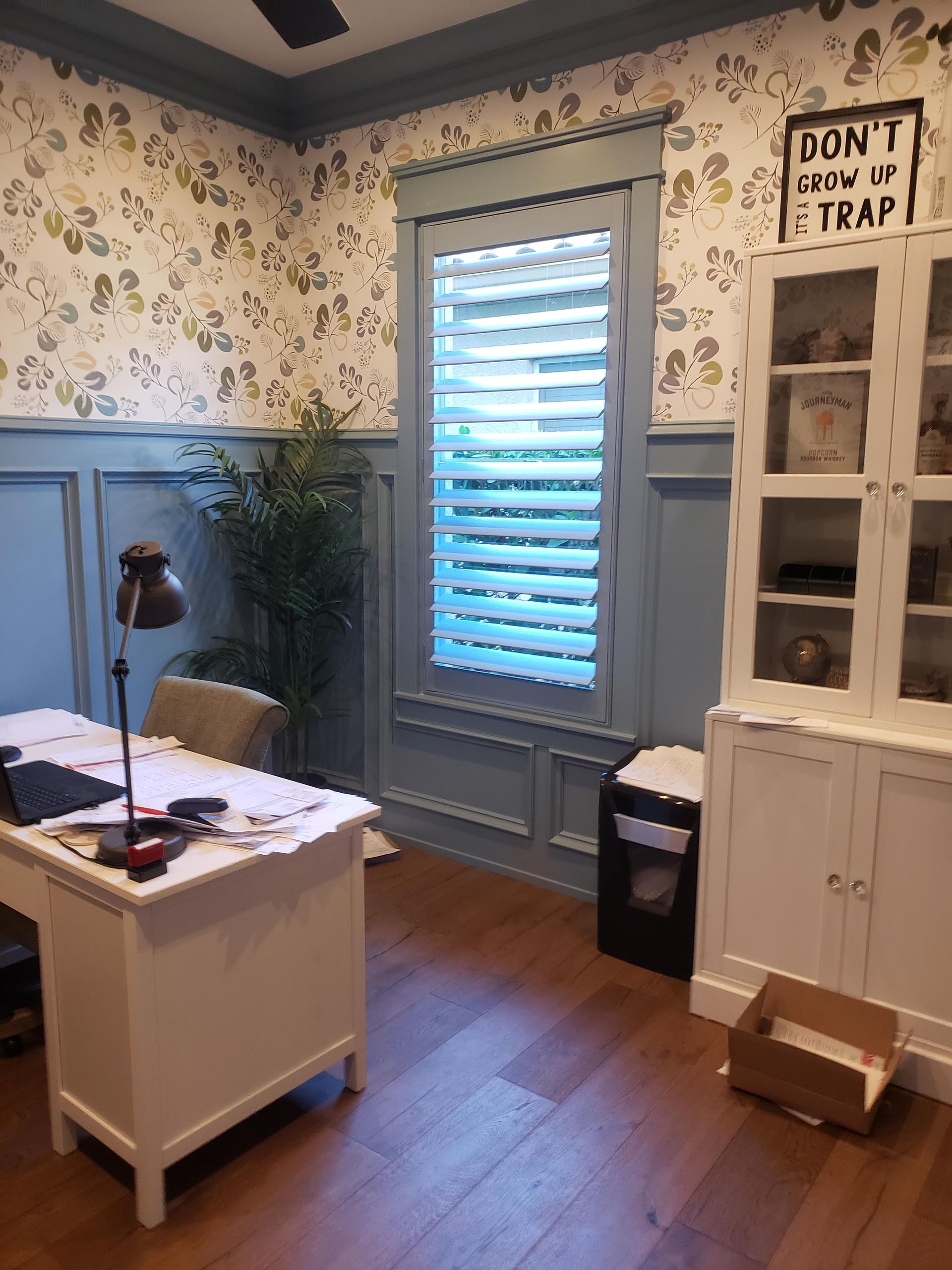 Office interior with white desk, blue paneled walls, floral wallpaper, large window with shutters, and white cabinet.