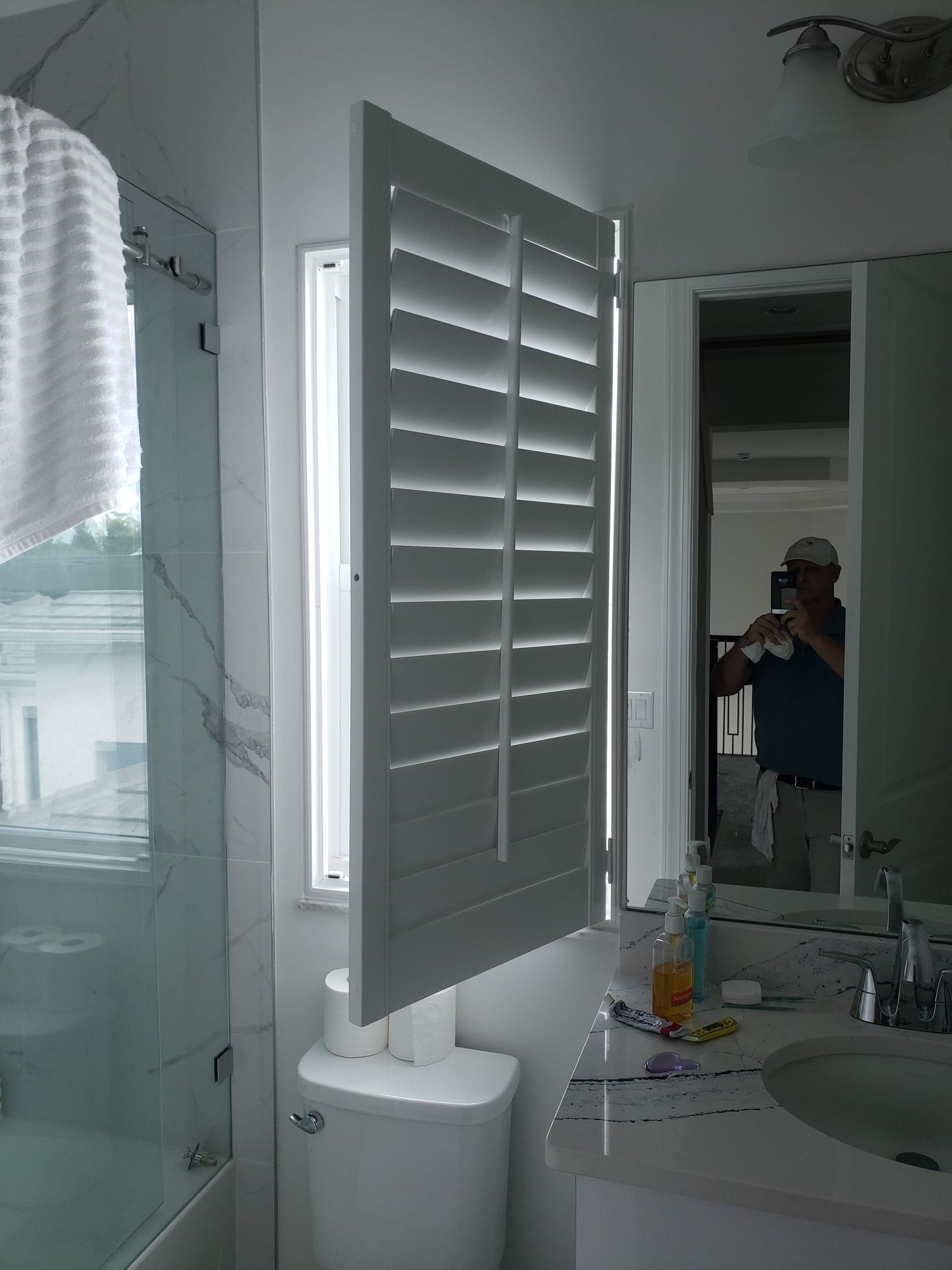 White bathroom shutters open over a toilet. A person is reflected in the mirror.