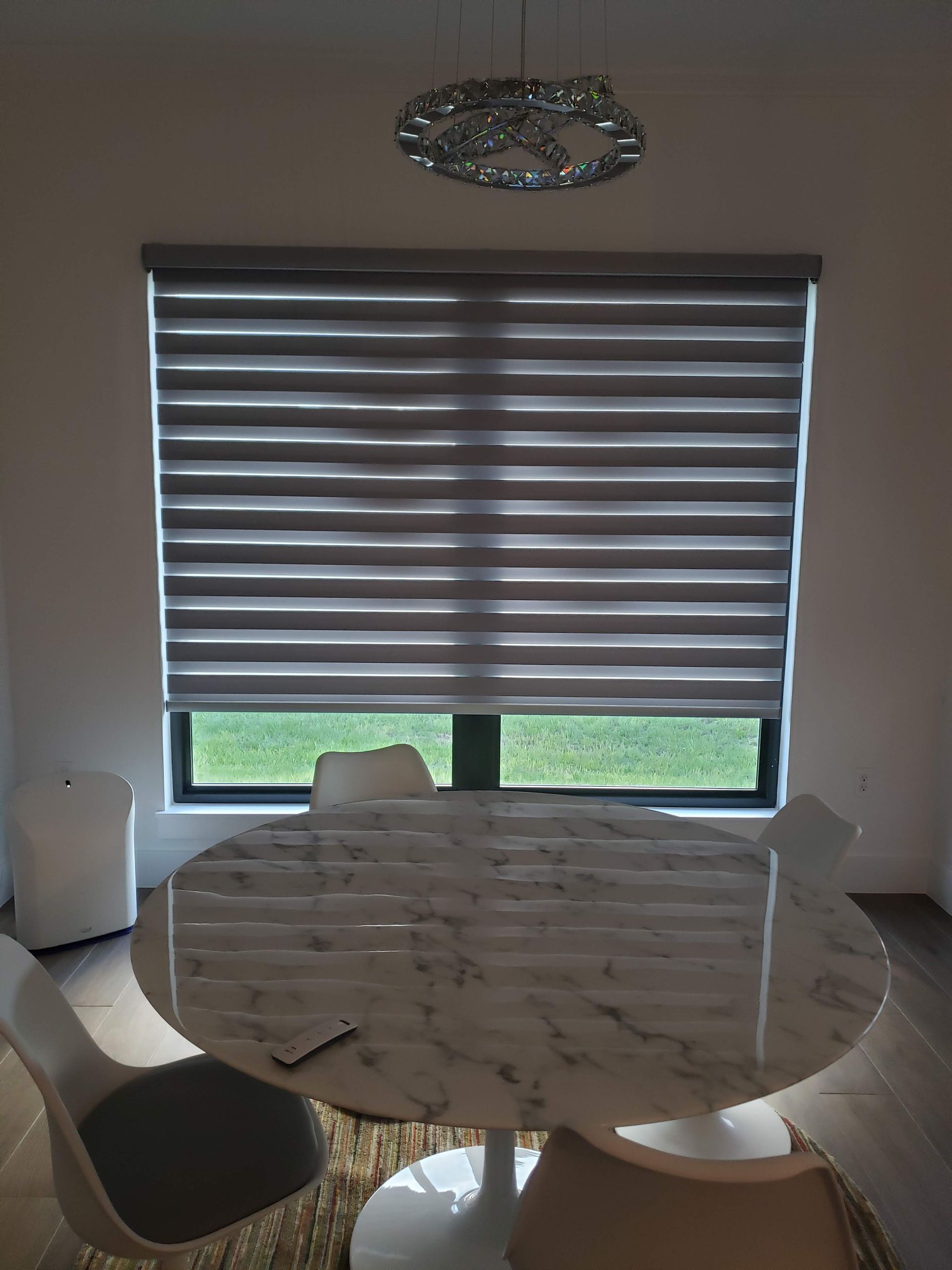 Dining room with window blinds, a marble table, and modern chairs. A chandelier hangs above.