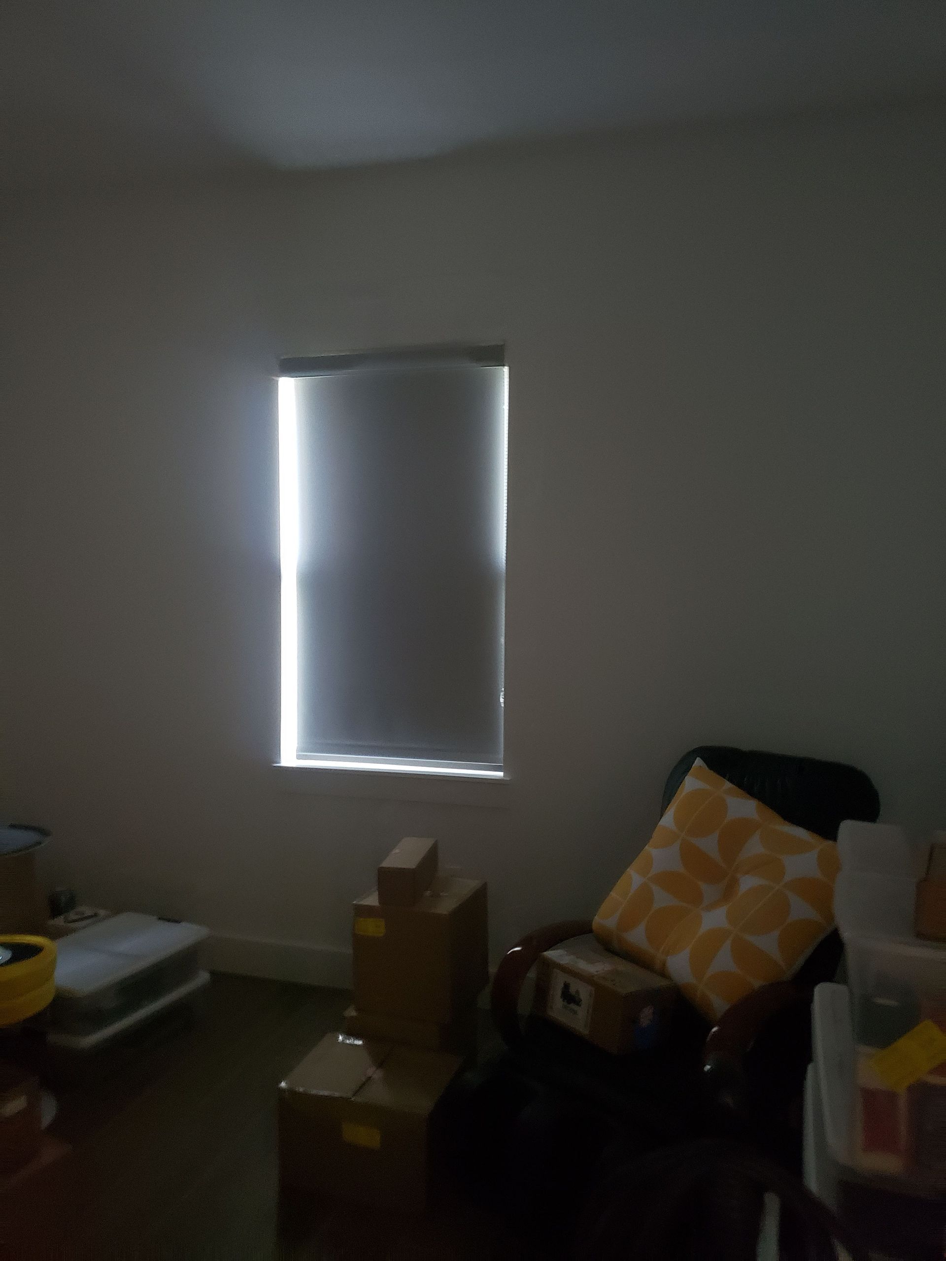 Window with a partially drawn shade lets in light, illuminating a mostly empty room with boxes, a chair, and a pillow.