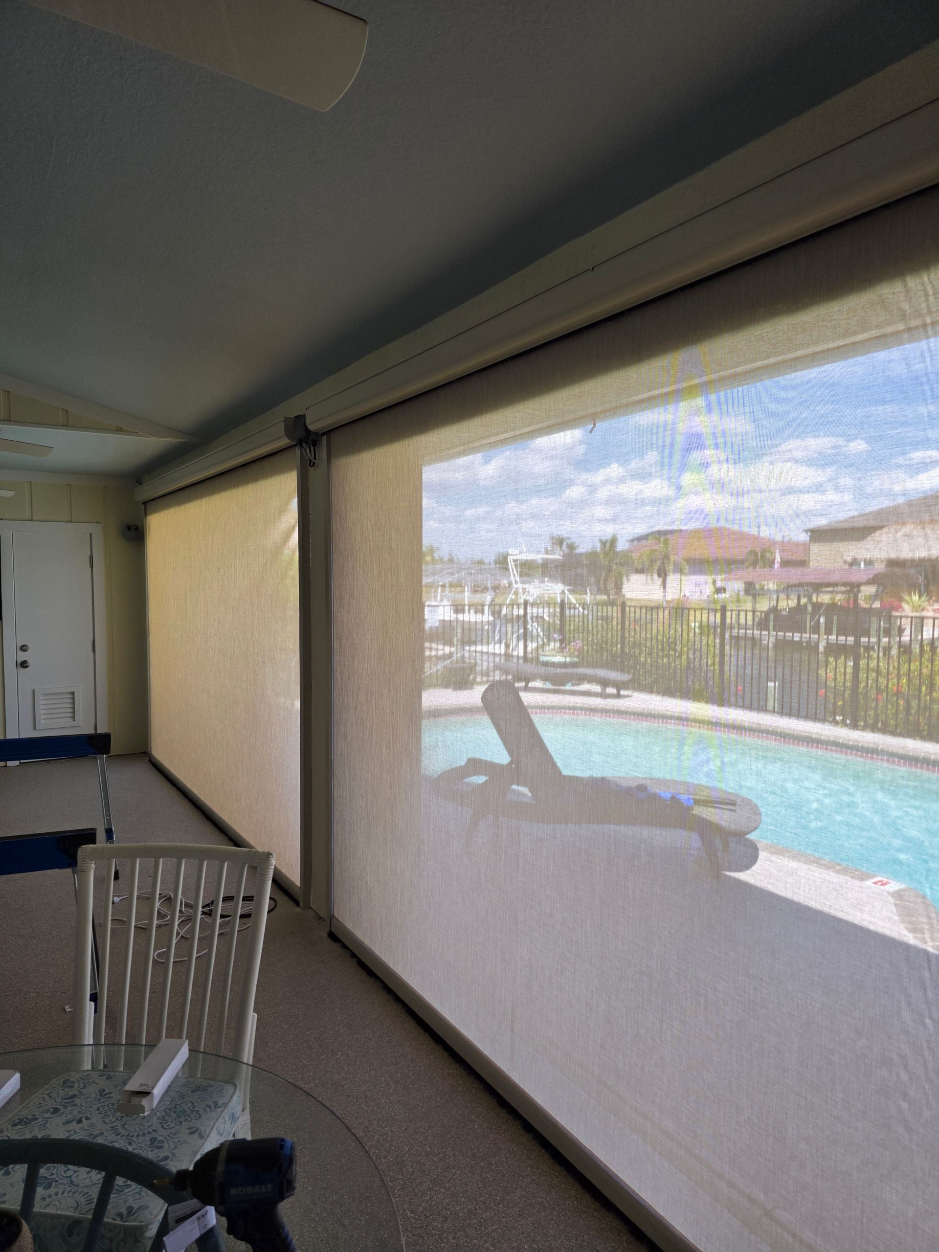 Patio with pool view, beige shade partially lowered, two white chairs, tan walls, and a glimpse of the outdoors.