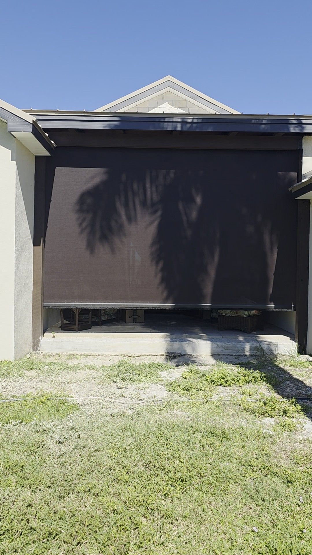 A dark brown sun shade partially lowered on a building, casting shadows onto the ground and a patch of grass.