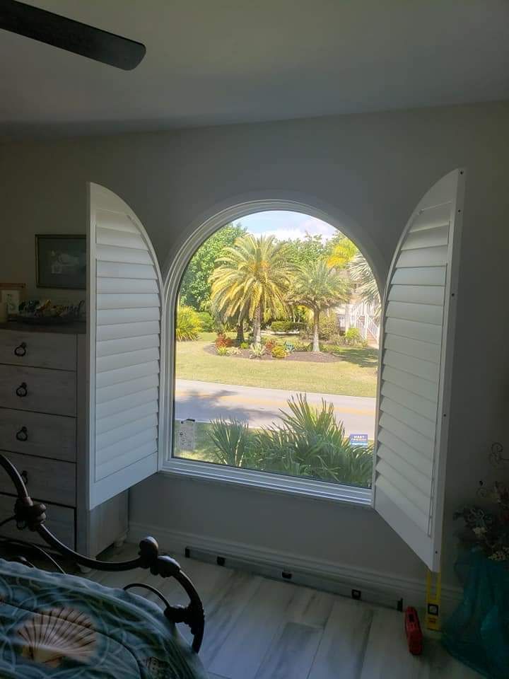 Arched window with white shutters open, revealing a sunny yard with palm trees.