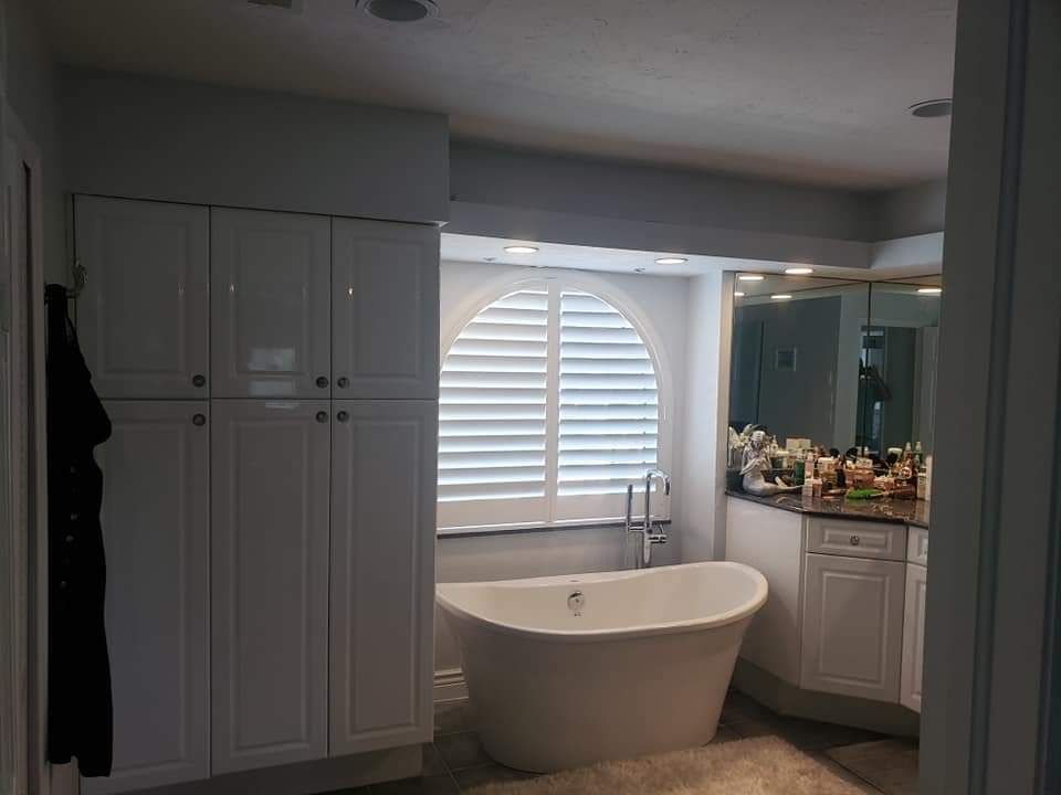 White bathroom with a freestanding tub, arched window with shutters, and white cabinetry.