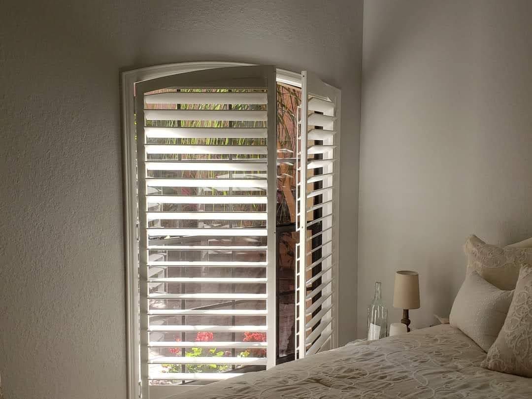 White shutters open over a window, with a bed in the corner.