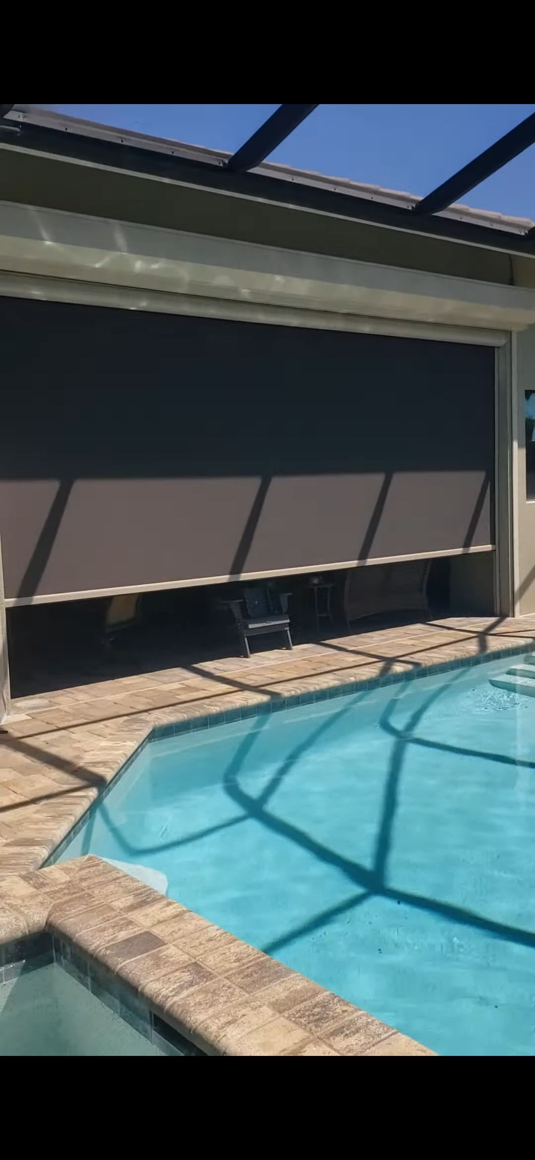 A pool next to a covered patio. A dark shade is lowered. Sunlight reflects off the pool.