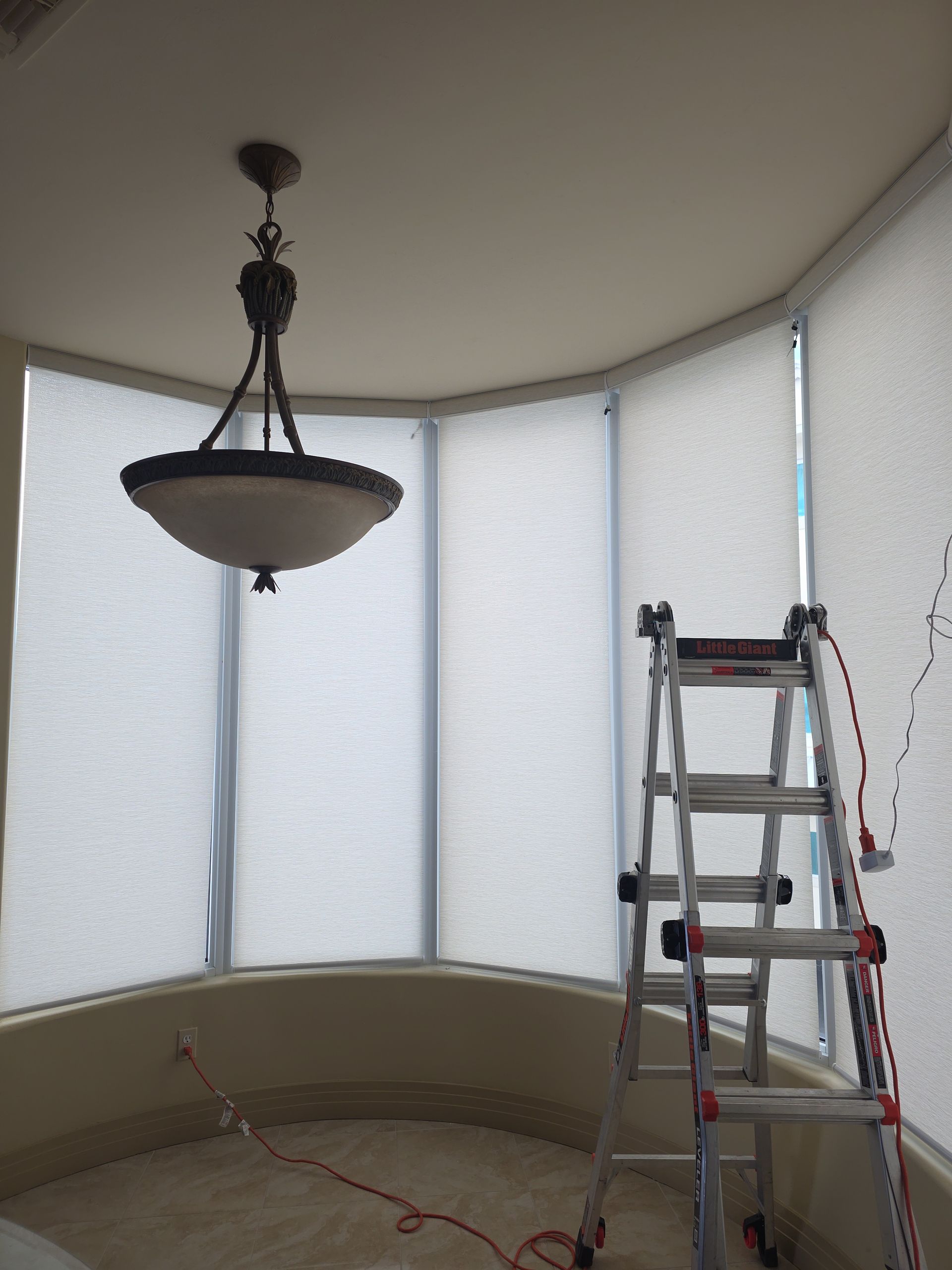 A chandelier hangs over a curved wall with white blinds. A ladder stands nearby.