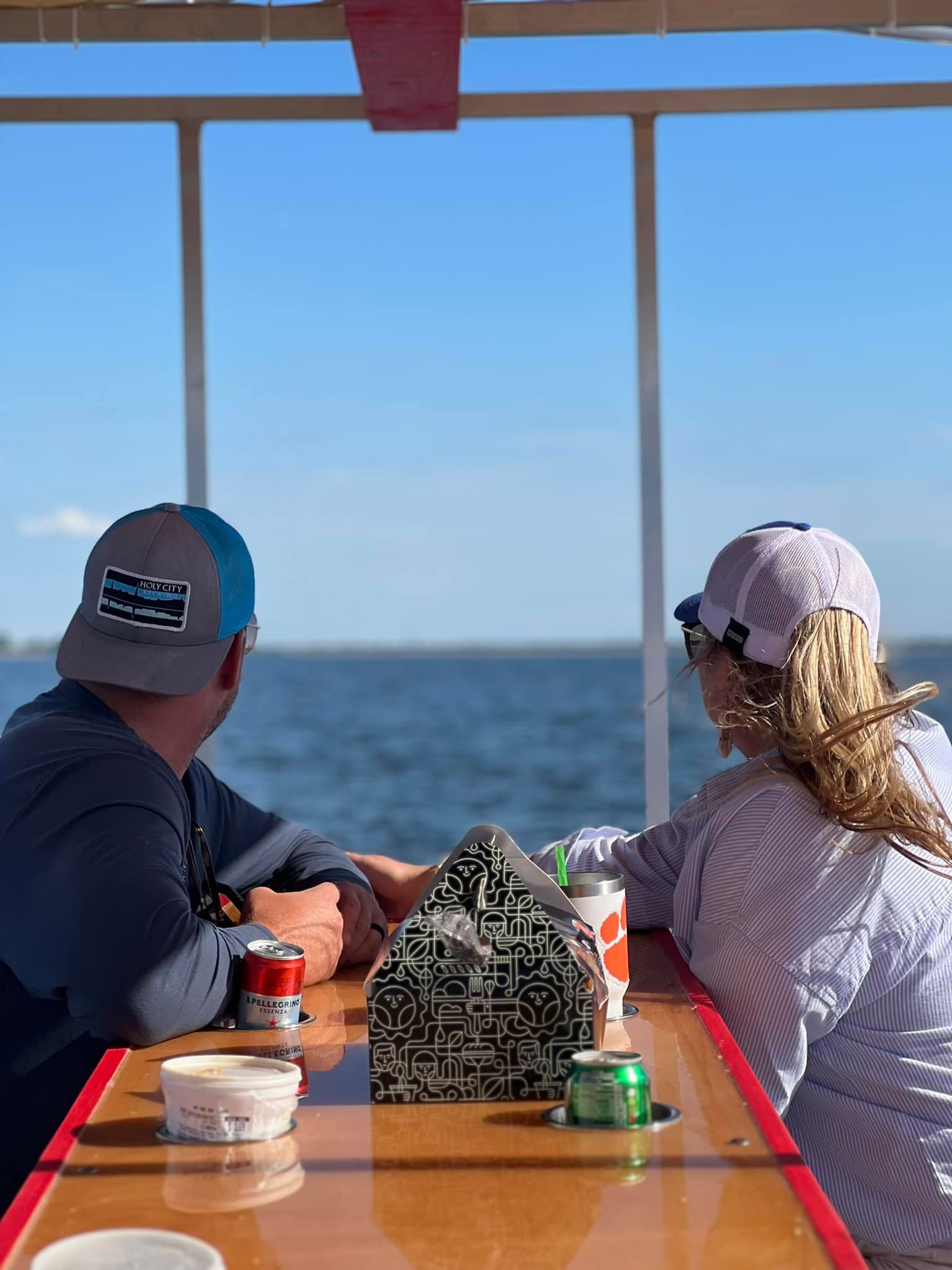 Two people sit at a table facing the water, wearing caps and looking out from a covered outdoor seating area.