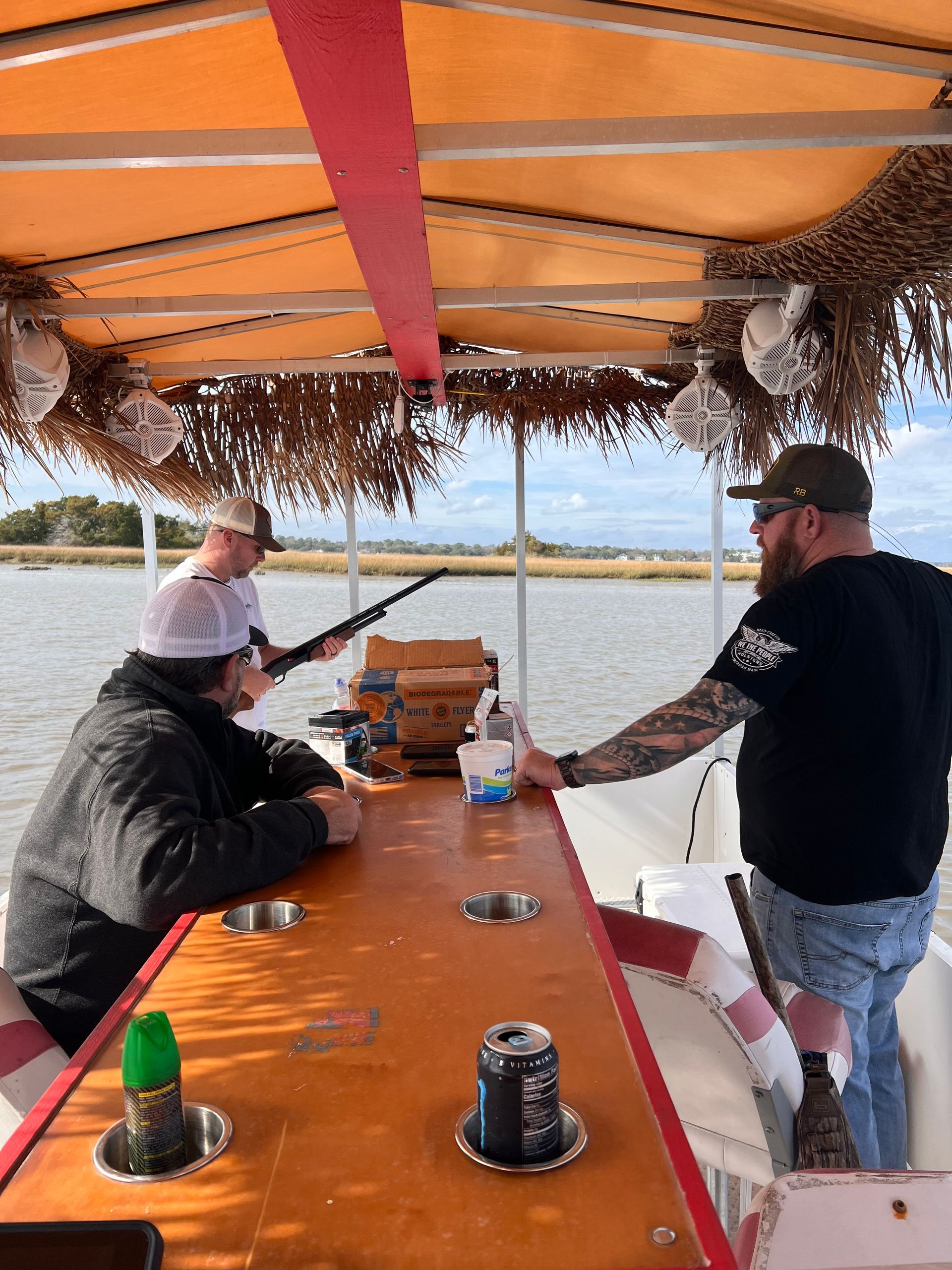 People on a boat under an orange canopy, fishing on calm water with rods and tackle on the table