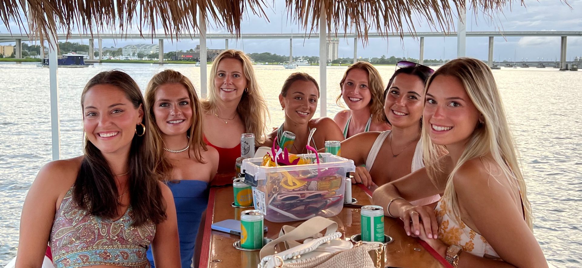 Group of friends smiling at a beachside table with drinks and a decorated cake under a thatched umbrella