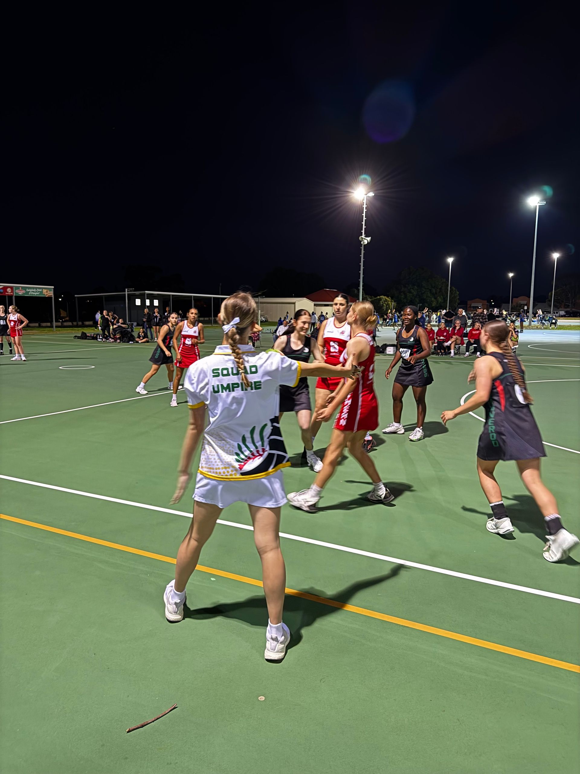 A group of women are playing a game of netball on a court at night.
