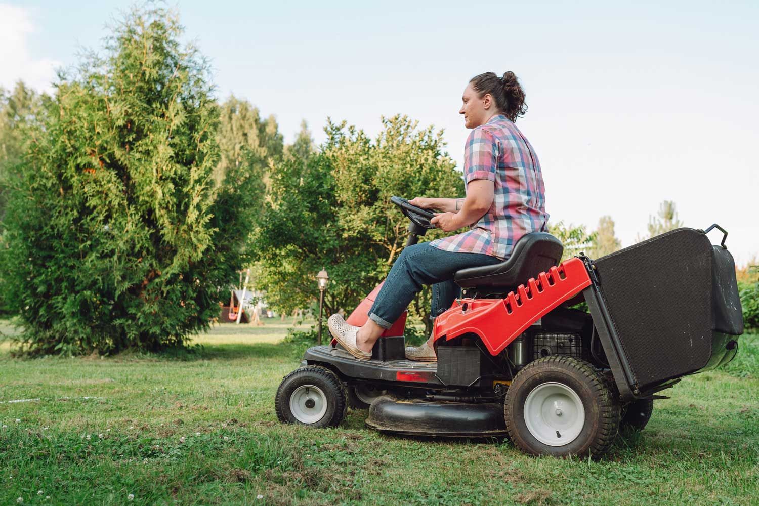 Person mowing a grassy yard with a red ride-on mower near trees and shrubs.