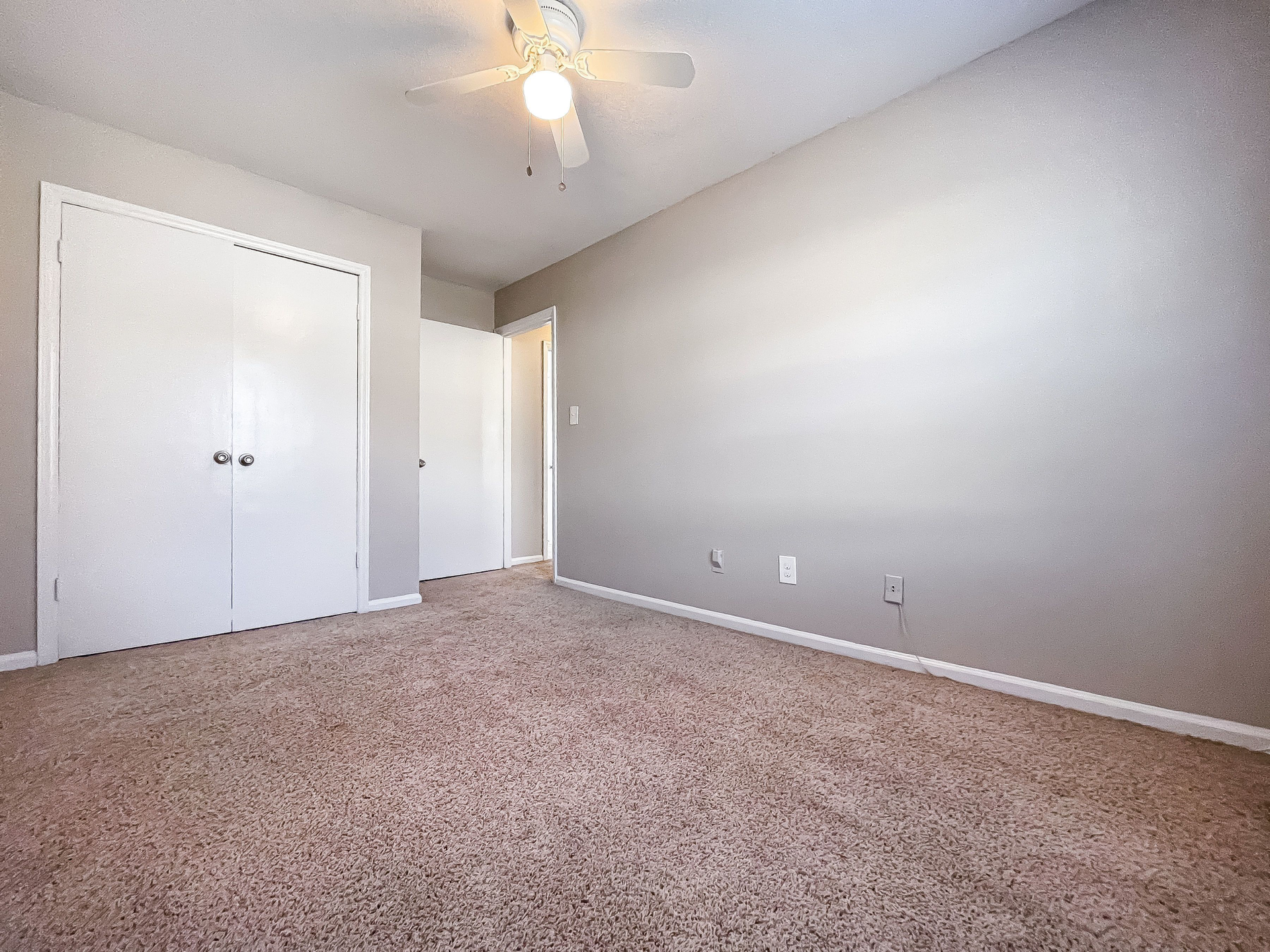 Bedroom interior with beige walls, carpet flooring, a ceiling fan, and double closet doors.