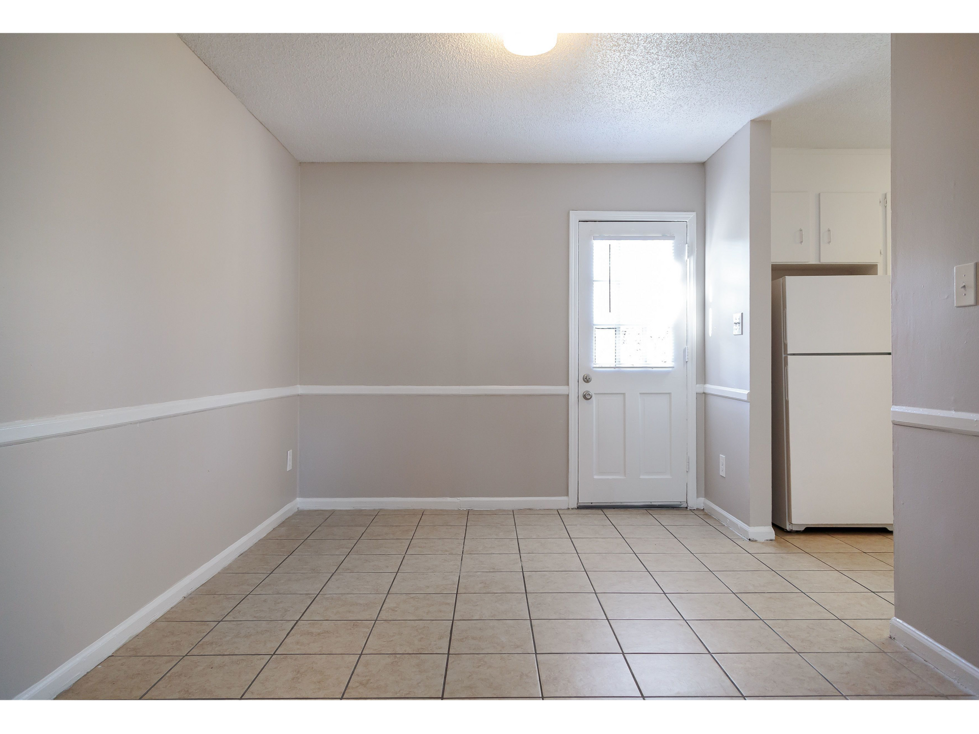Empty kitchen/dining area with tile floor, neutral walls, white exterior door, and a visible fridge.