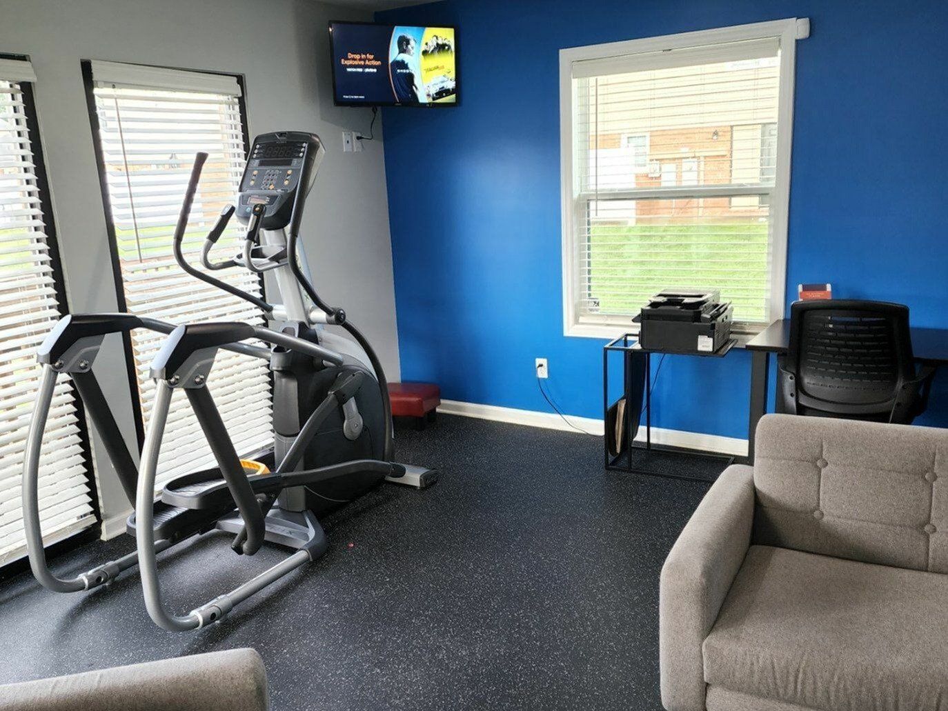 Compact on-site gym with cardio equipment, blue walls, windows, and a seating area.