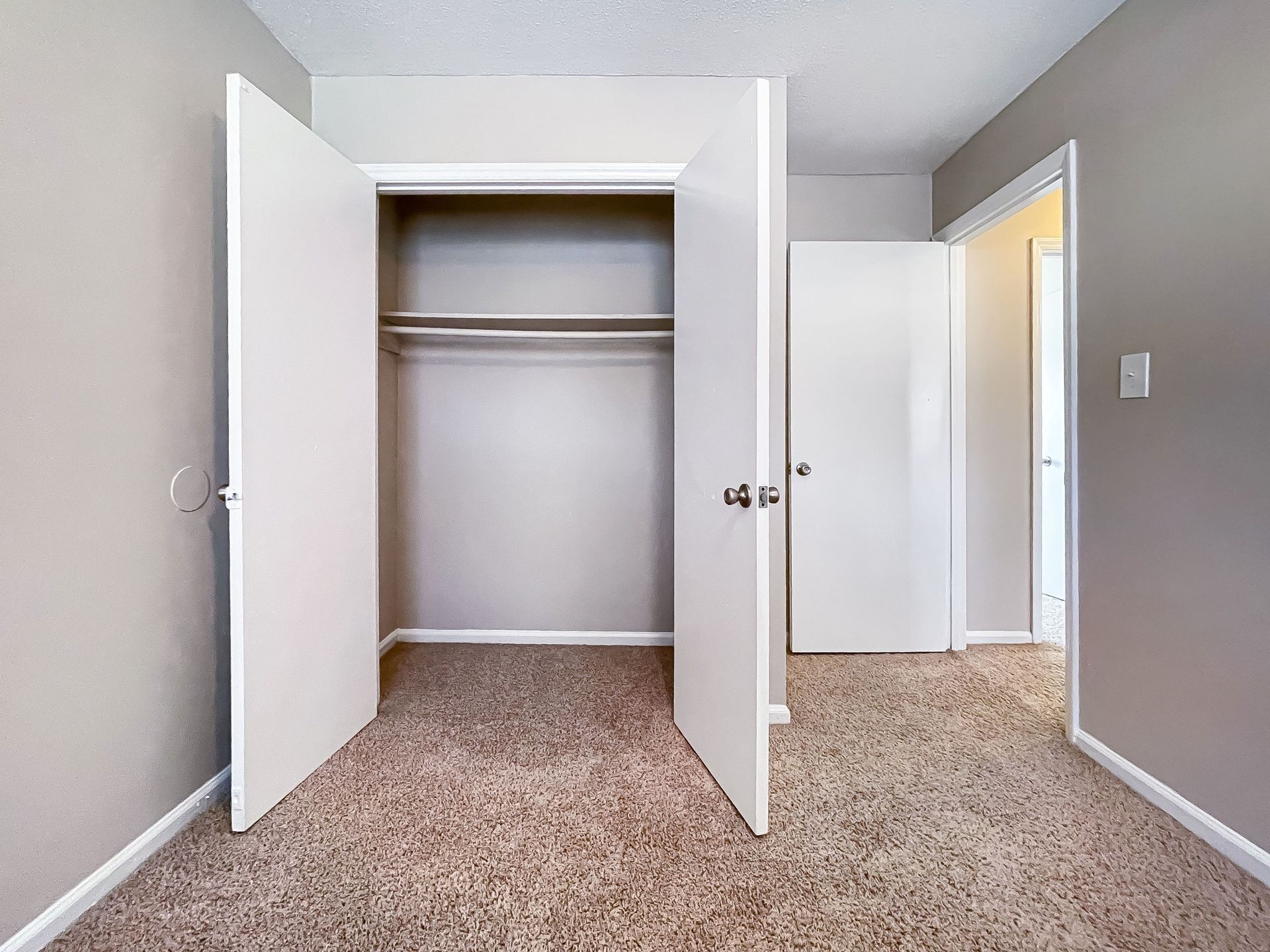 Bedroom with open double-door closet, hanging rod and shelf, beige walls and carpet.