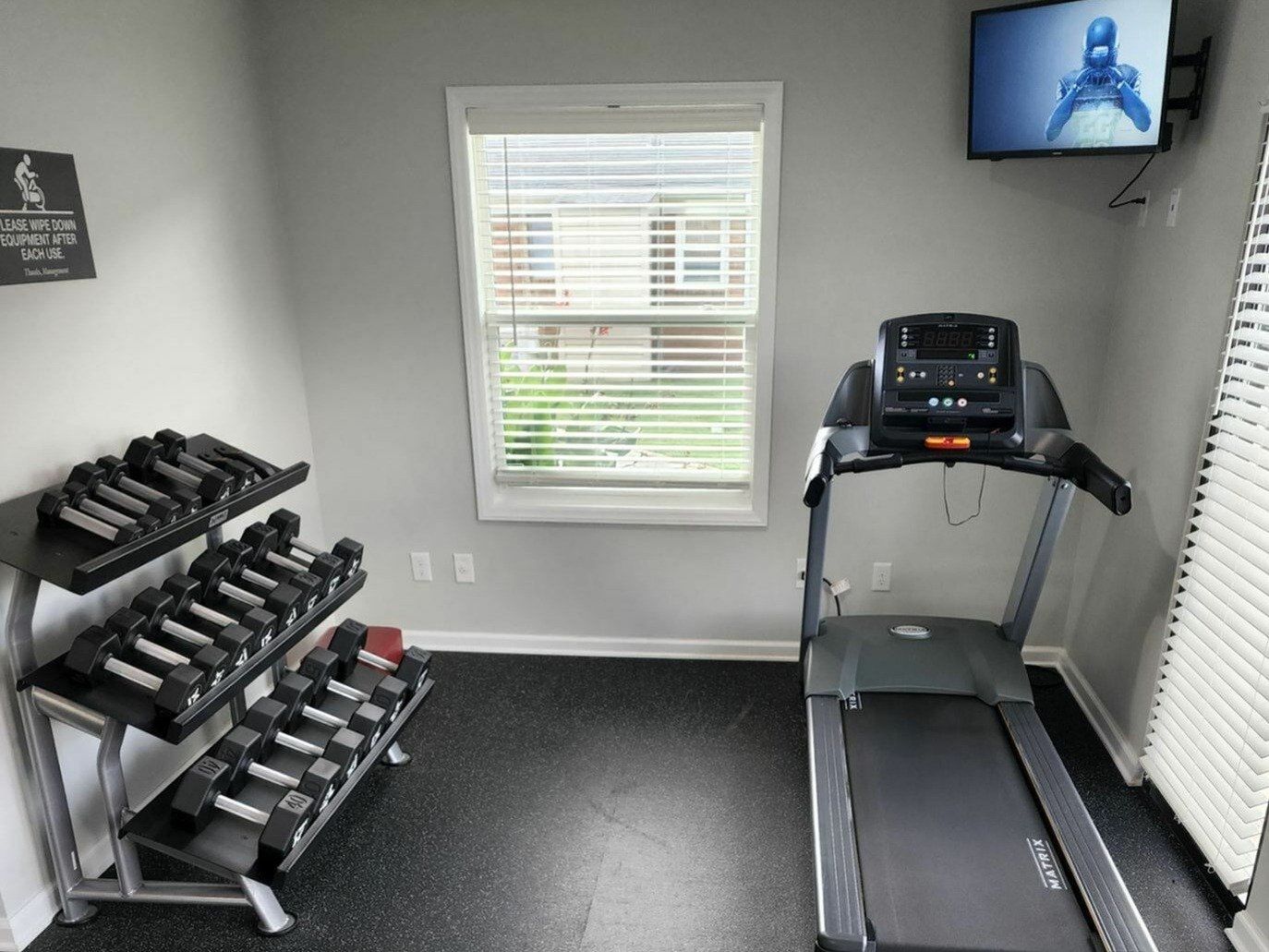 Small on-site gym with a treadmill and a rack of dumbbells by a window.